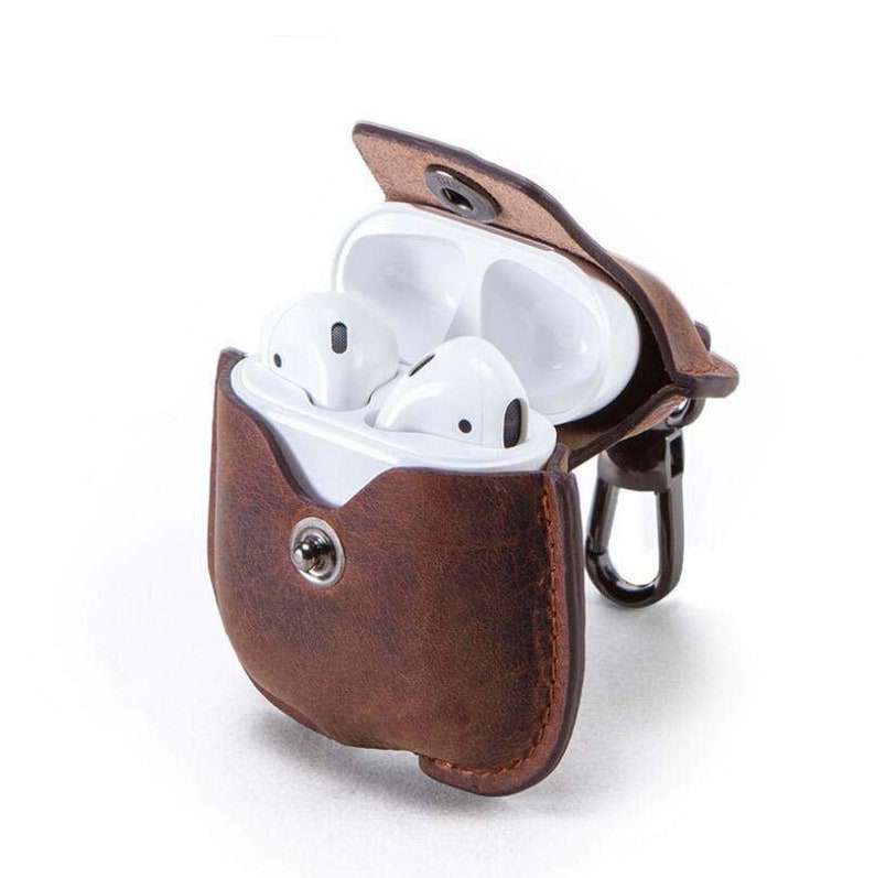 May include: A brown leather case with a snap closure for AirPods. The case has a metal carabiner attached to it.