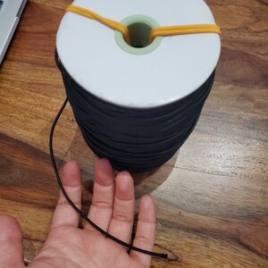 May include: A large spool of black elastic cord with a white core and a green center. The spool is secured with a yellow rubber band. A length of the black cord extends towards a hand.