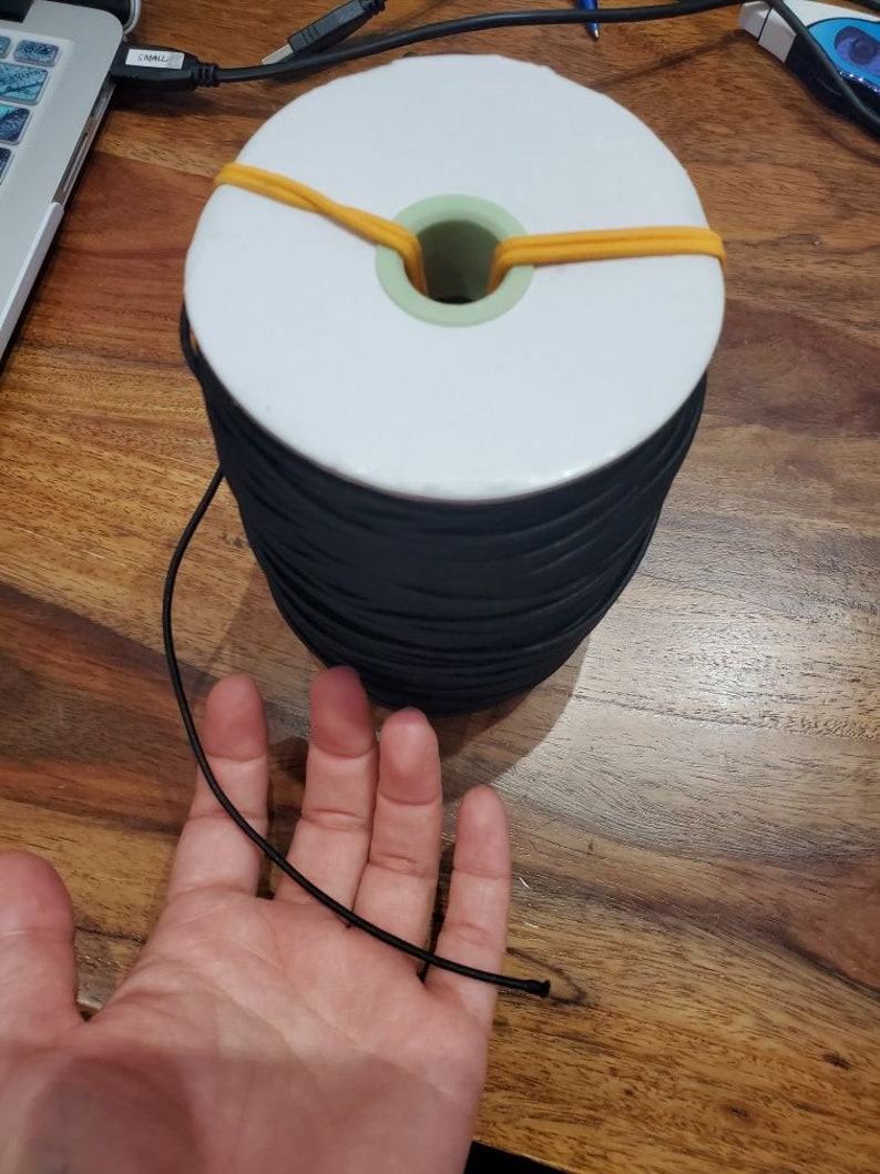 May include: A large spool of black elastic cord with a white core and a green center. The spool is secured with a yellow rubber band. A length of the black cord extends towards a hand.