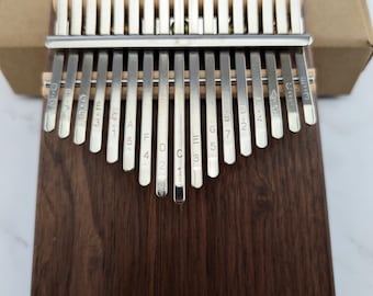 May include: A wooden kalimba with 17 metal tines, labeled with musical notes and numbers. The kalimba is made of dark brown wood and has a natural wood grain.