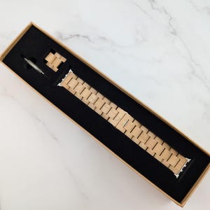 May include: A wooden watch band in a brown box. The band is made of light brown wood and has a silver buckle.