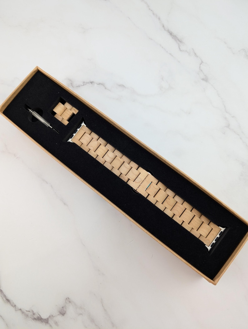 May include: A wooden watch band in a brown box. The band is made of light brown wood and has a silver buckle.