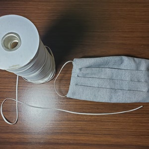 May include: A spool of white elastic cord with a gray fabric face mask. The elastic cord is wound around a white cardboard tube.