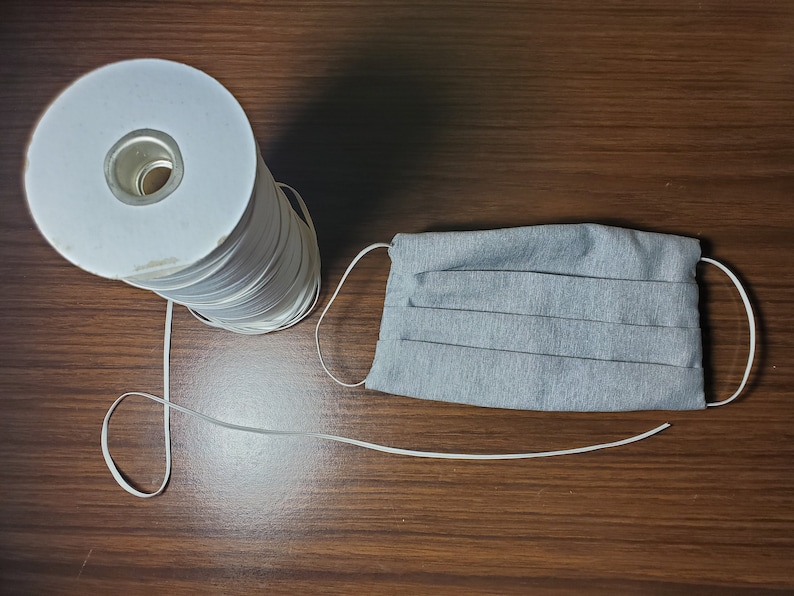 May include: A spool of white elastic cord with a gray fabric face mask. The elastic cord is wound around a white cardboard tube.