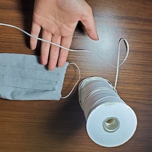May include: A spool of white elastic cord with a gray fabric face mask. The elastic cord is on a white cardboard spool.