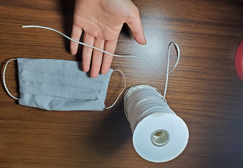 May include: A spool of white elastic cord with a gray fabric face mask. The elastic cord is on a white cardboard spool.