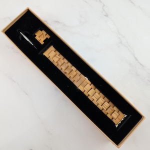 May include: A wooden watch band in a brown box. The band is made of light brown wood and has a silver buckle.