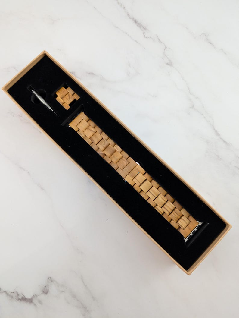 May include: A wooden watch band in a brown box. The band is made of light brown wood and has a silver buckle.