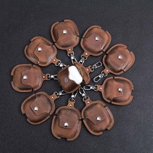 May include: Ten brown leather Airpod cases with a silver metal keychain attached to each. The cases are arranged in a circle with the keychains pointing outwards.