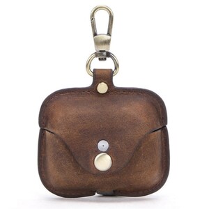 May include: A brown leather case with a gold metal carabiner clip for carrying AirPods. The case has a flap that closes with a snap.