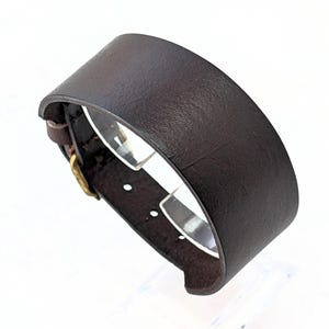Puede incluir: Una correa de reloj de cuero marrón oscuro con una hebilla dorada.