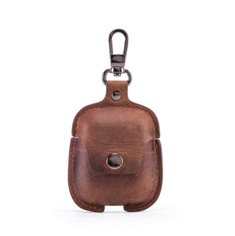 May include: A brown leather case with a metal carabiner clip for carrying AirPods. The case has a small flap with a button closure.