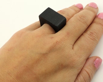 May include: A black, rectangular ring is worn on a finger. The ring has a solid, block-like design. The ring is made of a dark material and contrasts with the skin tone of the hand.