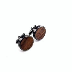 Puede incluir: Un par de gemelos negros con una incrustación redonda de madera. La madera tiene un color marrón oscuro y un patrón de veta natural.