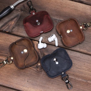 May include: Four leather cases for wireless earbuds with a metal keychain. The cases are in brown, dark brown, red, and black.