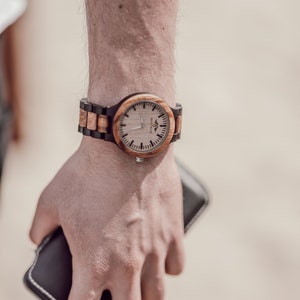 May include: A wooden wristwatch with a brown and black band. The watch face is white with black markings and a black logo.