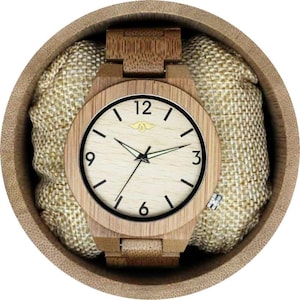 Può includere: Un orologio da polso in legno con un cinturino in bambù marrone chiaro e un quadrante color crema. Il quadrante dell'orologio ha lancette delle ore e dei minuti nere, indici delle ore in numeri romani neri e una lancetta dei secondi nera. L'orologio è in una scatola rotonda in legno marrone chiaro con una fodera in tessuto beige.