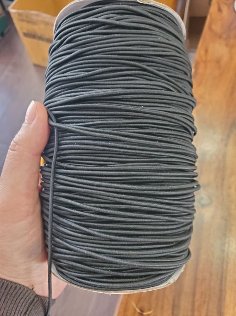 May include: A large spool of black elastic cord. The cord is tightly wound around a cardboard core. The elastic appears to be a solid black color and is held by a hand. The background is a wooden surface.