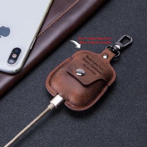 May include: Brown leather case with a metal carabiner clip for AirPods. The case has a pocket for a charging cable and a personalized inscription that reads "Bons Love You Merry Christmas 25.12.2020".