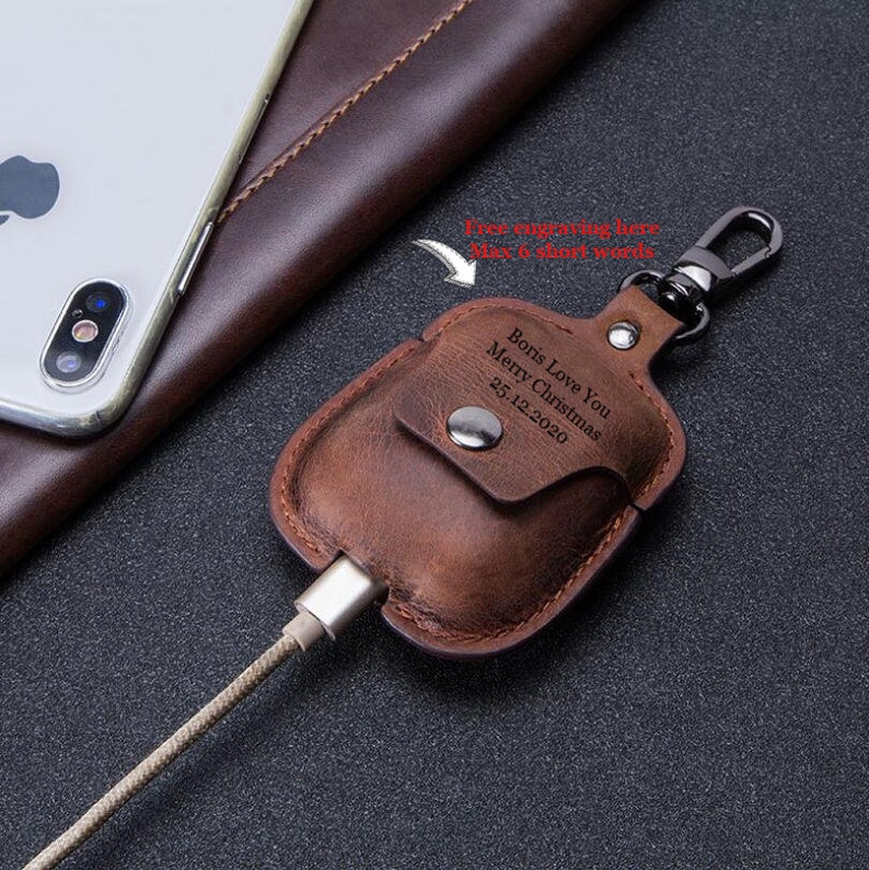 May include: Brown leather case with a metal carabiner clip for AirPods. The case has a pocket for a charging cable and a personalized inscription that reads "Bons Love You Merry Christmas 25.12.2020".