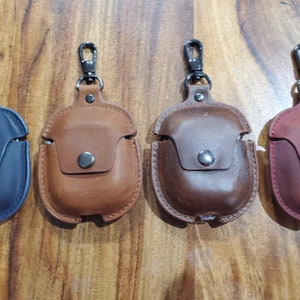 May include: Four leather Airpod cases with a metal carabiner clip. The cases are in navy blue, brown, dark brown, and burgundy.