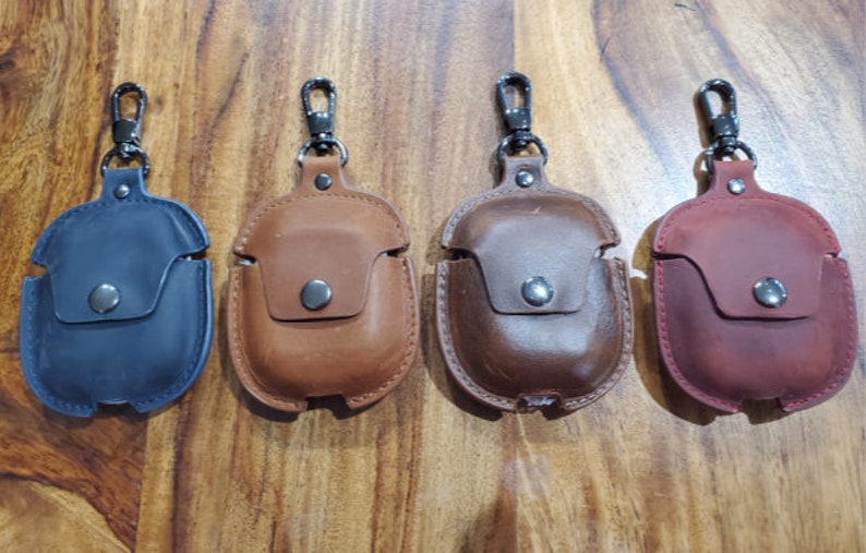May include: Four leather Airpod cases with a metal carabiner clip. The cases are in navy blue, brown, dark brown, and burgundy.