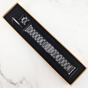May include: A gray metal Apple Watch band in a brown box with a black interior.