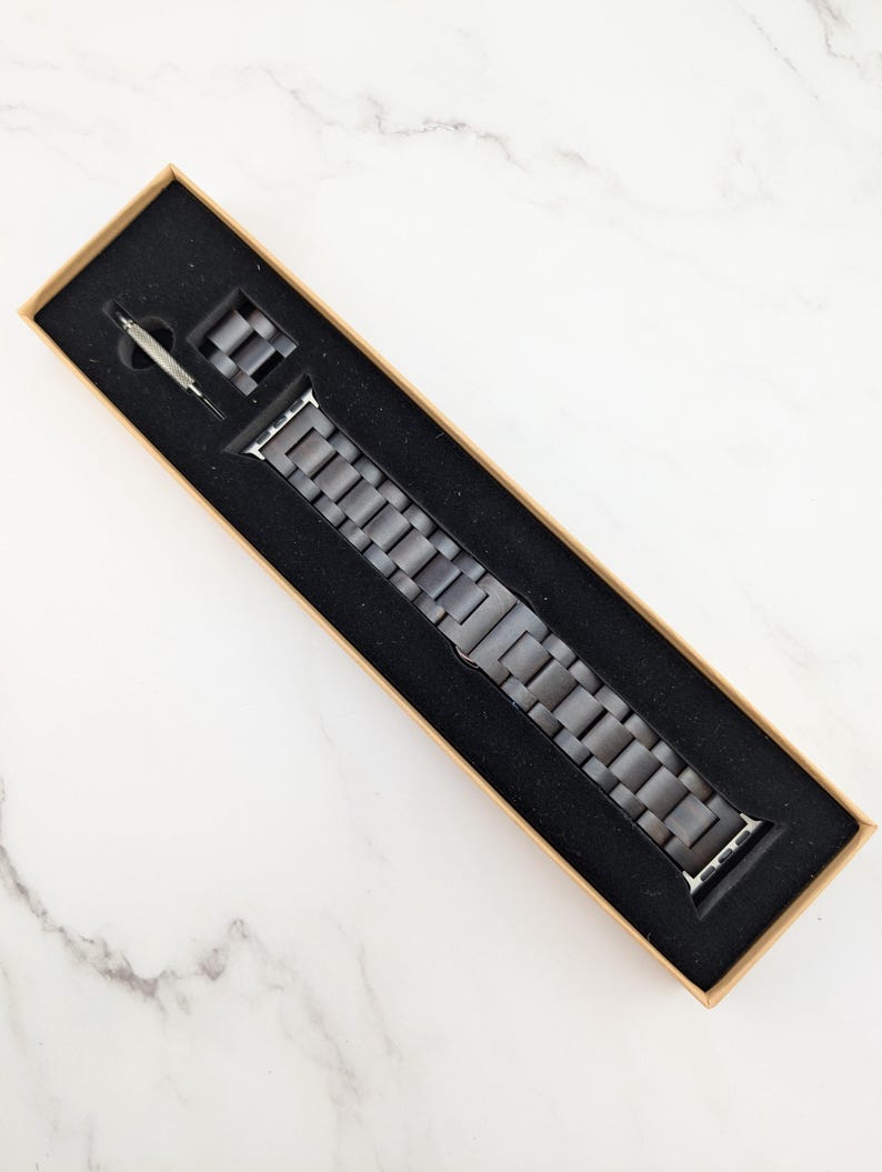 May include: A gray metal Apple Watch band in a brown box with a black interior.