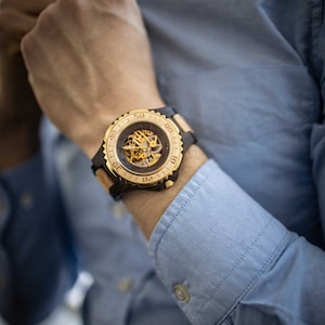 May include: A gold and brown wristwatch with a skeletonised dial and a black rubber strap. The watch has a rotating bezel with numbers.