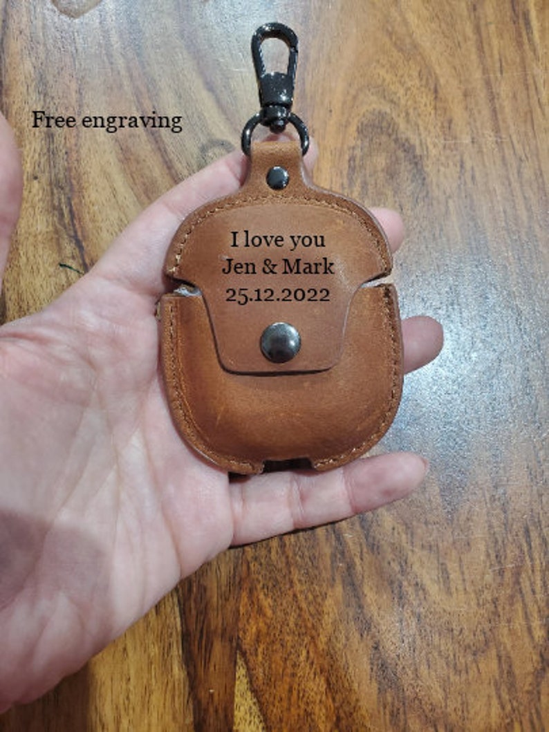May include: Brown leather Airpods case with a black metal carabiner clip. The case has the text "I love you Jen & Mark 25.12.2022" engraved on it.