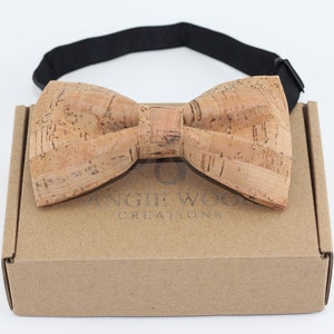 May include: A light brown cork bow tie with a black adjustable strap. The bow tie is in a brown cardboard box with the text "Angie Wood Creations" printed on the front.