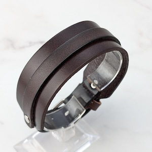 May include: A brown leather bracelet with a silver buckle. The bracelet is made of two layers of leather, and the buckle is a simple, rectangular design.
