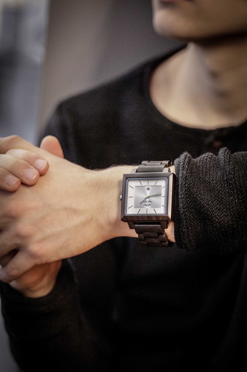 May include: A square-shaped wristwatch with a dark wood band and a white face. The watch has a silver-colored hour and minute hand and a small silver-colored dot at the 12 o'clock position.