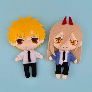 以下が含まれることがあります： 水色の背景に、2体のフェルト製キャラクター人形。1体は、とがった黄色の髪、白いシャツ、黒いネクタイ、黒いズボン。もう1体は、長いタン色の髪、赤い角、青いジャケット、黒いネクタイ、黒いズボン。人形には詳細な顔の特徴があります。