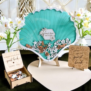 May include: A teal seashell-shaped guest book with a white frame. Features wooden seahorse and starfish cutouts, a wooden box, and two signs that read "Please sign our guest book". Ideal for a wedding.