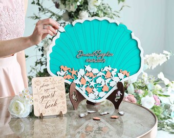 May include: A teal seashell shaped guest book with a white wooden stand. The shell is decorated with white and peach wooden sea creatures. The text "Daniel & Sophie 04.04.2024" is written on the shell. A sign that says "Please sign our guest book" is in the foreground.