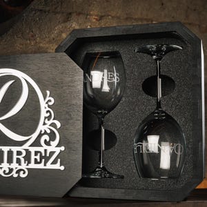 Puede incluir: Un juego de copas de vino personalizadas con dos copas, cada una grabada con un nombre, presentado en una caja de madera oscura. La caja tiene un diseño decorativo en la tapa. Las copas son transparentes con tallos largos.