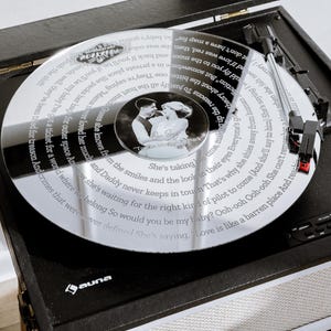 May include: A silver vinyl record with a photo and lyrics on a black turntable. The turntable is black with a silver speaker and control panel. The record player is on a white surface. The record player is a vintage style.