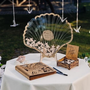 May include: A seashell-shaped guest book with a clear front and wooden cutouts of sea creatures. A wooden box with a sign that says "please sign" and a wooden guest book are on a white table. Decorative birds hang in the background.