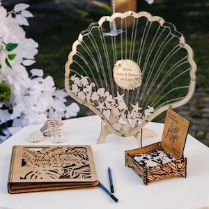 May include: A wooden seashell-shaped guest book alternative with a clear acrylic front, featuring wooden starfish and sea creatures. Includes a matching wooden box, a sign that says "please sign", and a wooden guest book. The names "Haley & Steven" and the date "July 15, 2026" are engraved.