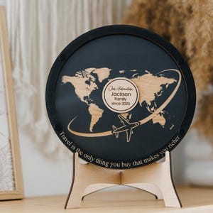 Può includere: Un cartello rotondo in legno nero a tema viaggio, con un design a mappa del mondo. Contiene il testo "Our Adventure Jackson Family since 2025" e "Travel is the only thing you buy that makes you richer".