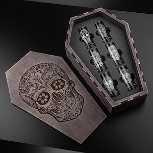 Può includere: Una scatola di legno a forma di bara con un teschio di zucchero decorativo inciso sul coperchio. All'interno, sei bicchierini con disegni di teschi e la scritta "Tequila Friends Forever". La scatola e i bicchieri sono scuri.