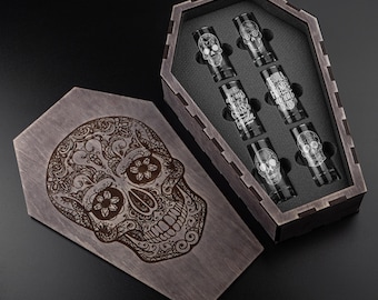 May include: A wooden coffin-shaped box with a decorative sugar skull engraved on the lid. Inside, six shot glasses with skull designs and the text "Tequila Friends Forever" are displayed. The box and glasses are dark-toned.