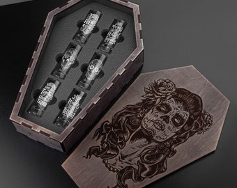 May include: A wooden coffin-shaped shot glass set with six glasses. The glasses have white text and skull designs. The coffin lid features a detailed engraved image of a Day of the Dead woman. The set is displayed on a black surface.