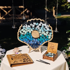 Puede incluir: Un libro de visitas en forma de concha de madera con recortes de criaturas marinas azules, turquesas y blancas. Incluye una caja de madera con un letrero que dice "please sign" y un libro de visitas de madera. Un recuerdo perfecto para bodas o eventos.
