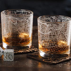 Può includere: Due bicchieri da whisky trasparenti incisi con un disegno di mappa della città, riempiti di liquido ambrato, poggiano su sottobicchieri decorativi. Accanto ci sono due cubi di pietra grigia con la lettera "R". I bicchieri e i cubi sono su una superficie scura.