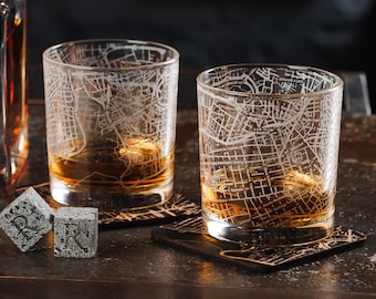 May include: Two clear whiskey glasses etched with a city map design, filled with amber liquid, sit on decorative coasters. Beside them are two gray stone cubes with the letter "R" etched on them. The glasses and cubes are on a dark surface.