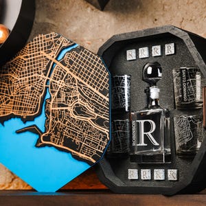 Puede incluir: Juego de decantador de madera negra con diseño de mapa de la ciudad. El juego incluye un decantador, vasos de chupito y una caja de madera. El decantador tiene una gran letra "R". La caja está abierta.