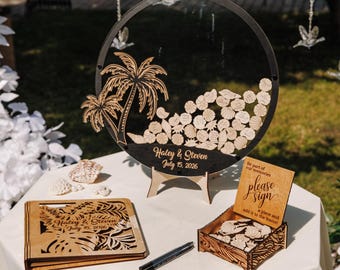 May include: A wooden wedding guest book set with a round sign-in board, a wooden box, and a guest book. The round sign-in board has a black background with a palm tree design and the names "Haley & Steven" and the date "July 15, 2026".