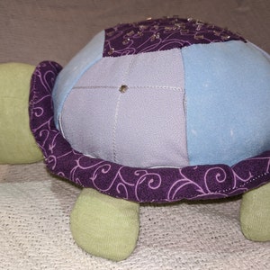 May include: A patchwork fabric turtle toy with a purple and blue shell and green legs. The shell is decorated with a purple swirl pattern.